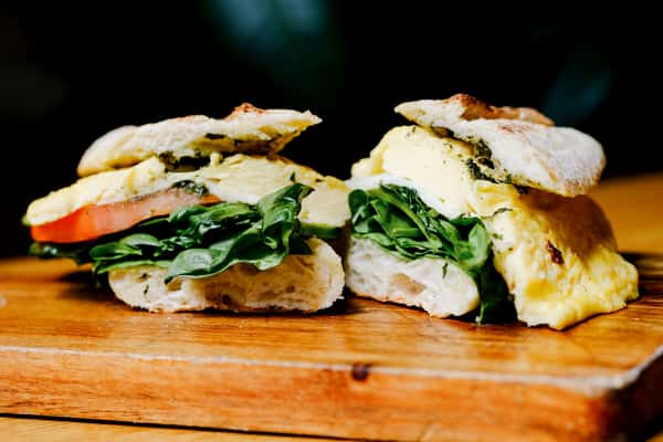 Gourmet breakfast sandwich on rustic ciabatta, layered with herb egg, spinach, and tomato, served on a wooden board in a warm café setting.