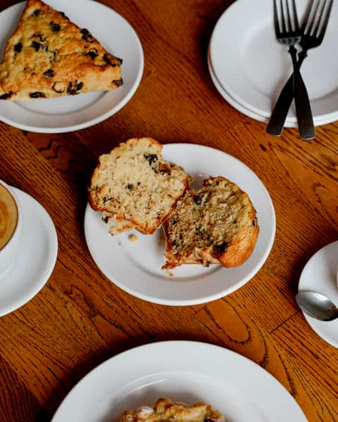 Top-down view of a sliced raisin muffin on a white plate, served with a golden scone, lattes, and extra cutlery on a wooden café table.