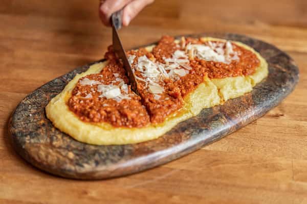 Hand cutting into a serving of creamy Italian polenta topped with rich Bolognese meat sauce and shaved Parmesan cheese, presented on a rustic stone platter over a wooden table.