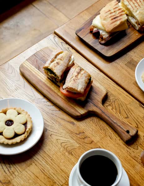 Toasted ciabatta sandwiches with tomato, mozzarella, and pesto on wooden boards, paired with a jam-filled flower tart and black coffee on a rustic café table.