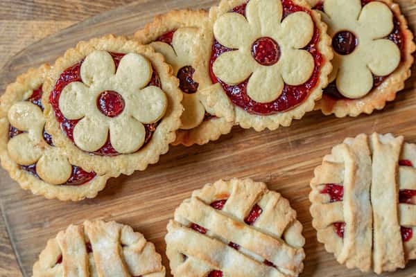 Close-up of decorative jam-filled cookies featuring flower and lattice tops, arranged on a wooden tray for a festive, handmade bakery presentation.