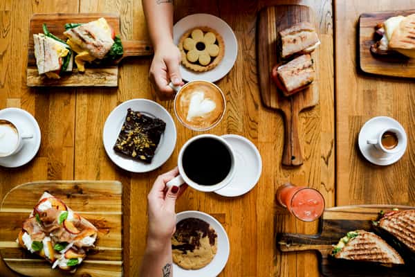 Colorful brunch spread featuring specialty coffee drinks, cookies, brownies, and gourmet breakfast sandwiches, laid out on a rustic wooden café table.