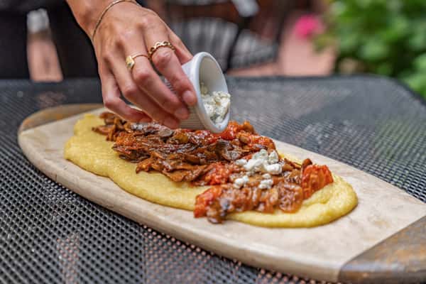 Creamy polenta topped with savory braised meat and tomato sauce, being garnished with crumbled cheese by hand, presented on a rustic wooden board — a hearty Italian comfort dish.