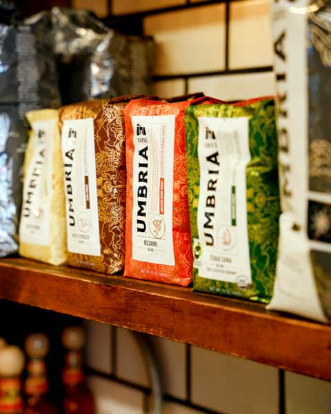 Colorful lineup of Caffè Umbria whole bean coffee bags neatly displayed on a rustic wooden shelf, showcasing a variety of artisanal espresso blends.