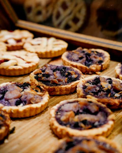Close-up of freshly baked mini tarts with mixed berry and nut toppings, arranged on a wooden tray inside a bakery display case, showcasing golden crusts and vibrant fruit fillings.