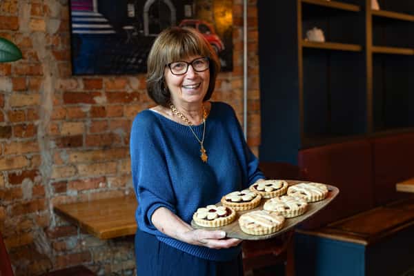 Smiling woman holding a tray of handmade jam-filled cookies with flower and lattice designs, standing inside a cozy bakery with exposed brick and wooden décor.