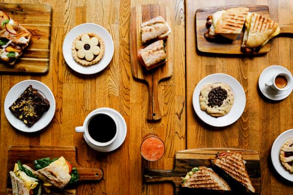 Top-down view of a rustic café brunch spread with sandwiches, cookies, brownies, espresso shots, and fresh juice on wooden boards and plates.