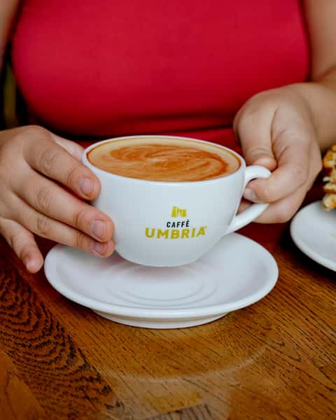 Close-up of a creamy latte in a Caffè Umbria-branded cup, held with both hands over a wooden café table, highlighting warm tones and artisanal coffee presentation.