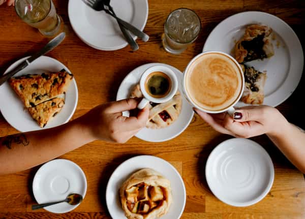 Café brunch moment with two people clinking an espresso and a latte over a table filled with pastries and iced drinks, capturing a warm, social vibe.