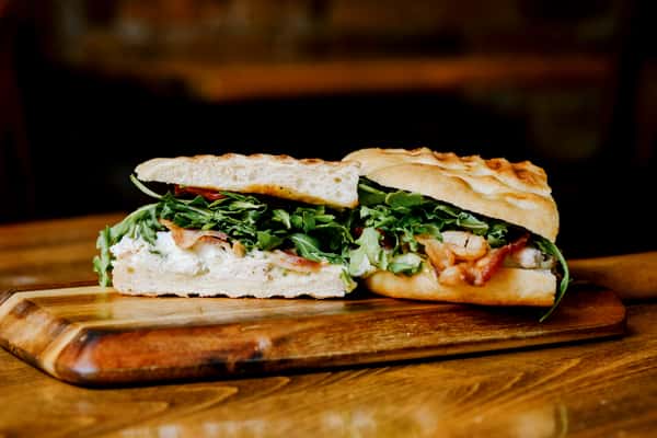 Close-up of an artisan focaccia sandwich filled with arugula, creamy spread, crispy bacon, and fresh tomato, served on a wooden board in a cozy café setting.