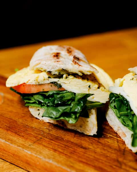 Close-up of a gourmet breakfast sandwich on ciabatta bread, layered with fluffy herb-seasoned eggs, fresh spinach, and ripe tomato slices, served on a rustic wooden board — perfect for a hearty café brunch.