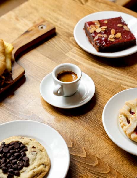 Rustic café table with a single espresso shot surrounded by a chocolate chip cookie, almond-topped brownie with fruit glaze, jam-filled lattice pastry, and a grilled breakfast sandwich on a wooden board — perfect for a cozy brunch or coffee break.