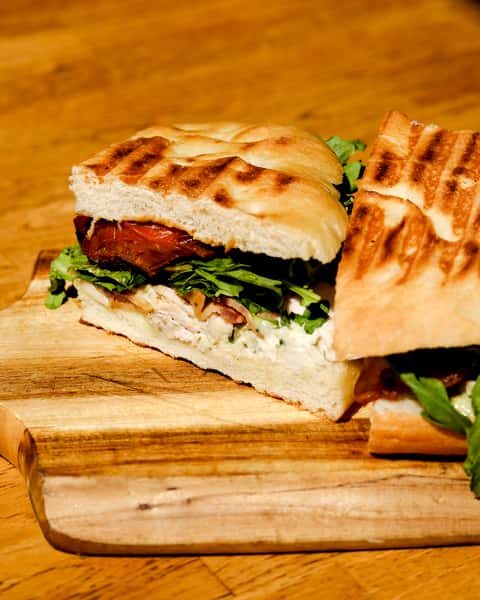 Grilled focaccia sandwich filled with arugula, roasted tomato, creamy spread, and savory chicken, served on a wooden board.