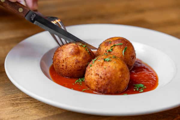 Close-up of three golden-brown arancini being cut into, served over a vibrant tomato sauce and garnished with fresh herbs—classic Italian appetizer on a white plate over a wooden table.