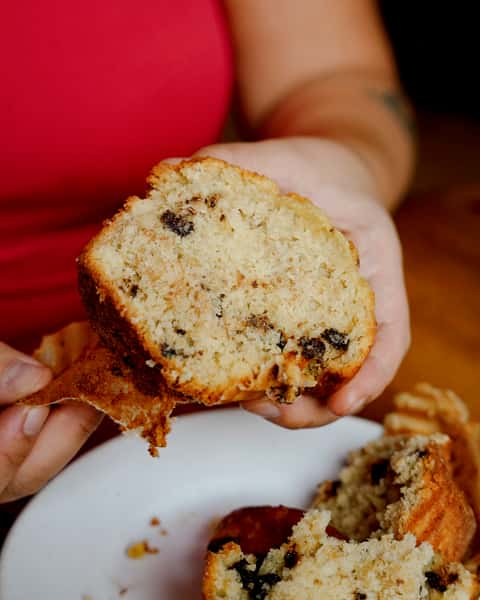 Close-up of a hand holding a moist chocolate chip muffin split open, revealing its fluffy texture and rich filling on a white plate.