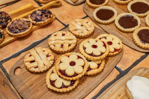 Selection of decorative jam tarts with lattice and flower tops alongside chocolate-filled tartlets, artfully arranged on wooden trays for a bakery or dessert buffet.