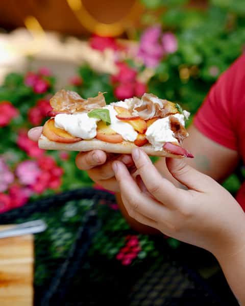 Hand holding an open-face sandwich topped with fresh peach slices, prosciutto, whipped ricotta, and basil, against a garden backdrop.