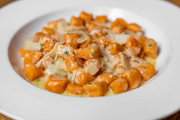 Close-up of creamy pasta with shaved parmesan, mushrooms, and sausage in a white wine sauce, served in a white plate on a wooden table.