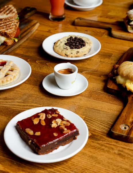 Artisan café breakfast spread on a rustic wooden table, featuring a rich chocolate brownie topped with almond slices, a single shot of espresso, a large chocolate chip cookie, a strawberry-glazed pastry, grilled paninis, and a breakfast sandwich with eggs and cheese — perfect for a cozy brunch atmosphere.