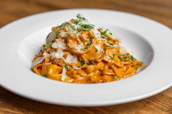 Tagliatelle pasta coated in a rich, creamy tomato sauce with crumbled sausage, topped with shaved Parmesan and fresh parsley—classic Italian comfort food served on a white plate.