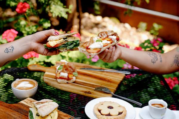 Two people clinking gourmet open-face breakfast sandwiches topped with prosciutto, peach slices, and ricotta over a vibrant garden café table.