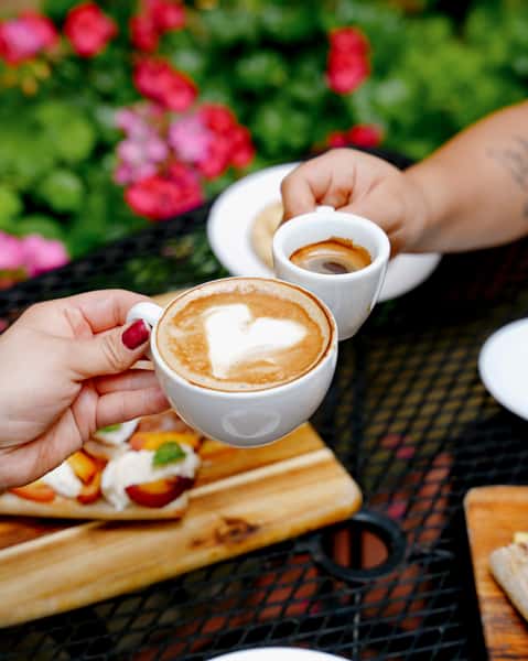 Hands toasting with a cappuccino and an espresso in an outdoor café garden setting, surrounded by floral décor and gourmet breakfast boards.