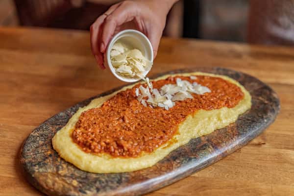 Creamy polenta spread topped with rich meat ragu, being finished with fresh parmesan shavings — a traditional Northern Italian dish served on a rustic stone platter.