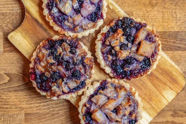 Overhead shot of four baked tarts with a rich filling of dark berries and diced pear, served on a wooden paddle board—perfect for artisan bakeries or seasonal dessert spreads.