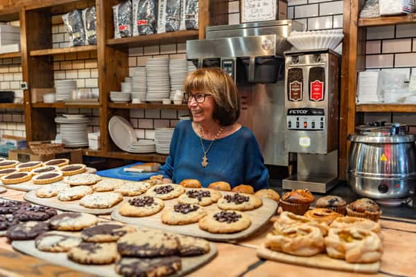 Smiling woman behind a bakery counter filled with fresh-baked cookies, muffins, pastries, and tartlets, set in a cozy café with rustic wood shelving, white subway tiles, and commercial coffee equipment.