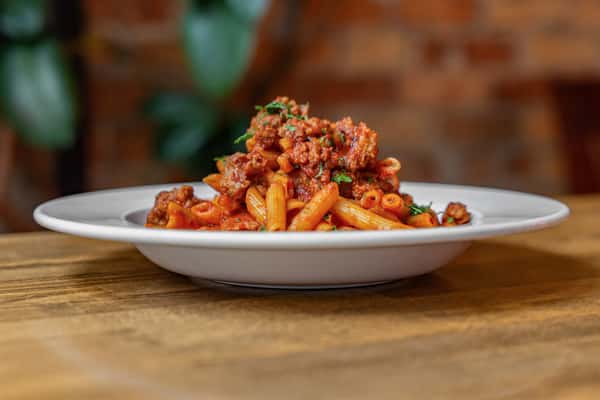 Penne pasta served with hearty Italian sausage ragù and fresh herbs, presented in a shallow white bowl over a rustic wood surface—perfect for a traditional Italian main course.