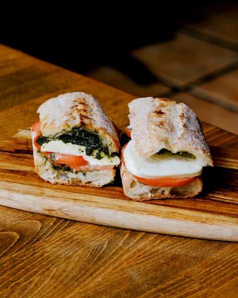 Italian-style caprese sandwich with fresh mozzarella, tomato, and basil pesto on rustic ciabatta bread, served on a wooden board.