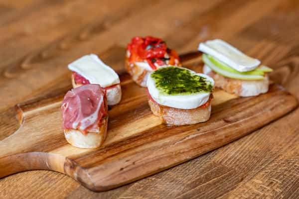 Assortment of gourmet Italian crostini on a rustic wooden board, topped with fresh mozzarella and pesto, roasted red peppers, prosciutto, brie with jam, and brie with green apple—ideal for an antipasto platter or appetizer menu.