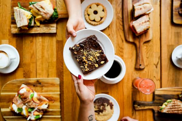 Overhead view of a hand passing a rich chocolate brownie topped with crushed nuts, with various cookies, sandwiches, and coffee drinks in the background.