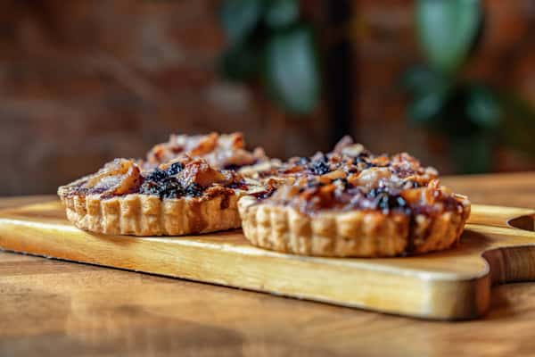 Side view of artisanal fruit tarts topped with a glossy blend of dark berries and diced pear, displayed on a wooden serving board against a rustic backdrop.