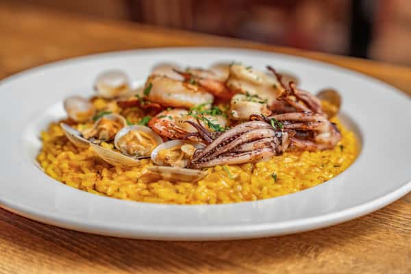 Close-up of saffron risotto topped with grilled shrimp, calamari, and clams, garnished with herbs—classic Italian seafood risotto served in a white bowl on a rustic wood table.