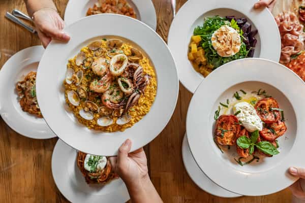 Top-down view of a vibrant Italian seafood risotto plated with shrimp, clams, and calamari, surrounded by fresh Mediterranean dishes including burrata with grilled tomatoes, a beet and mango salad, and pasta entrées, all served on white plates over a wooden table.