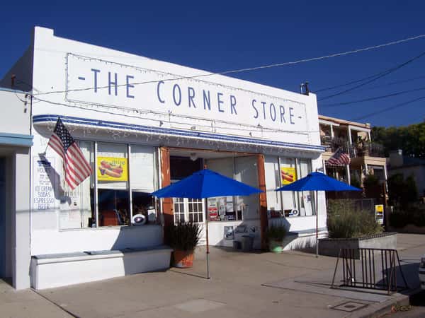 Exterior of The Corner Store café with white storefront, blue signage, American flags, and outdoor umbrellas, capturing a classic neighborhood café vibe on a sunny day.