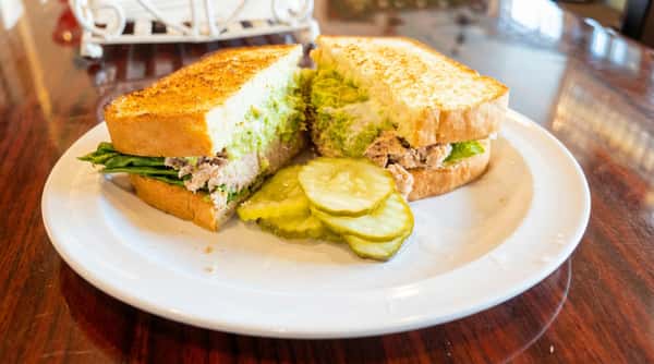 Tuna salad sandwich on toasted bread, with visible green avocado spread, lettuce, and sliced pickles.