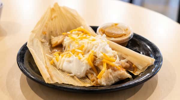 Tamales on a black plate, topped with shredded cheddar-jack cheese and a dollop of sour cream, with a side of salsa.