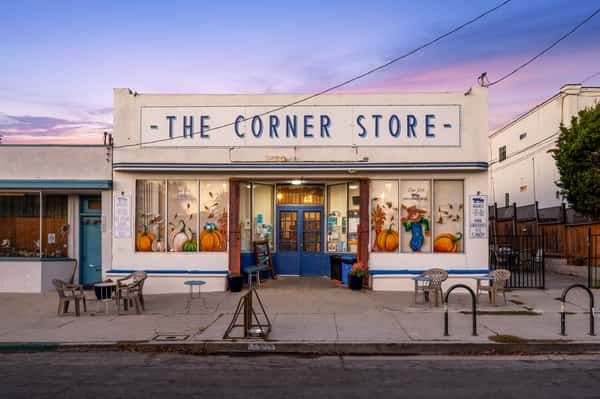 The Corner Store café exterior at sunset with blue doors, seasonal window art, and sidewalk seating, showcasing a cozy local café and market in a charming neighborhood setting.