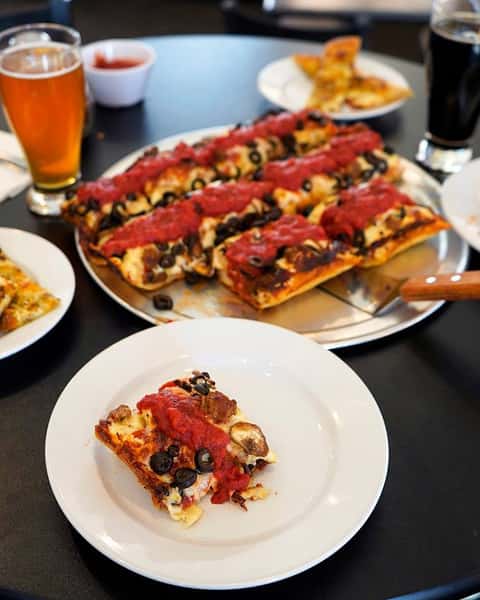 square pizza topped with mushrooms, olives and marinara sauce on a table with a glass of beer