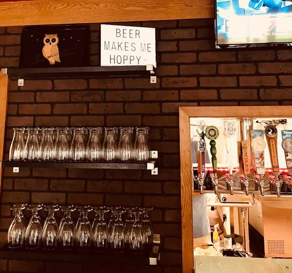 brick wall with a window to the kitchen. Beer taps located inside the window. Shelves on the wall with assorted glasses. Top shelf holds a sign that says "beer makes me hoppy".