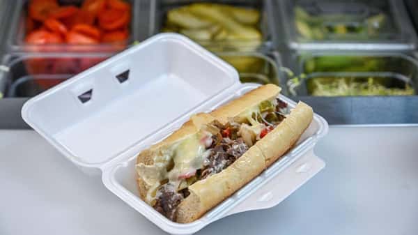 A fresh Philly cheesesteak sandwich in a to-go container, loaded with thinly sliced beef, melted cheese, sautéed onions, and peppers. Fresh sandwich toppings like tomatoes, pickles, and shredded lettuce are visible in the prep station in the background.