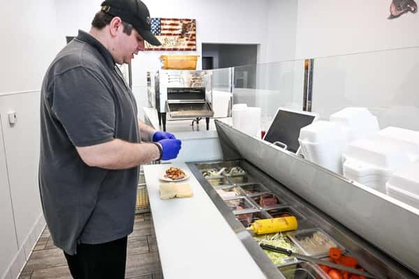 A sandwich maker at Eagle Sandwich Co. prepares a fresh sub behind the counter, wearing gloves for food safety. An assortment of deli meats, cheeses, vegetables, and condiments are neatly organized in a refrigerated prep station.