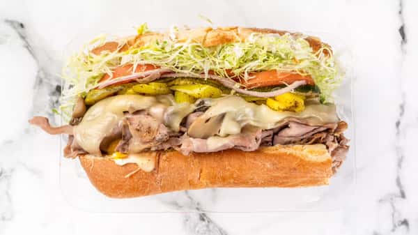 Hearty roast beef sub sandwich with melted provolone cheese, banana peppers, sliced pickles, tomatoes, red onions, and shredded lettuce on a toasted hoagie roll, served in a clear takeout container.