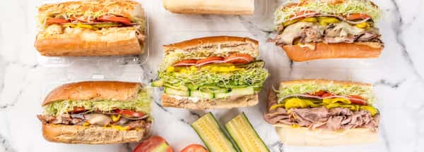 Assorted gourmet sandwiches from Eagle Sandwich Co., filled with fresh deli meats, melted cheese, crisp lettuce, tomatoes, pickles, and cucumbers, served on toasted hoagie rolls with a side of cucumber slices.