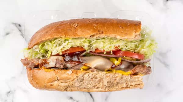 Hearty roast beef sub sandwich with melted cheese, tangy barbecue sauce, shredded lettuce, tomato slices, and red onion on a toasted wheat hoagie roll, served in a clear takeout container.