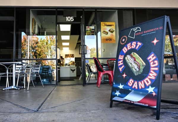 Exterior of Eagle Sandwich Co. with a bold sign reading "The Best Sandwich," featuring a sandwich image, outdoor seating with metal tables and chairs, and an open door revealing the counter inside.