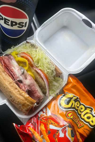 Hearty deli sandwich with melted cheese, sliced salami, banana peppers, fresh tomatoes, red onions, and shredded lettuce on toasted bread, served in a takeout box with a bag of Cheetos and a Pepsi drink.