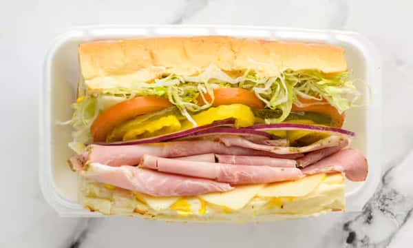 Classic sub sandwich with layers of ham and Swiss cheese, topped with shredded lettuce, tomato slices, banana peppers, pickles, and red onion on a fresh hoagie roll, served in a takeout container.